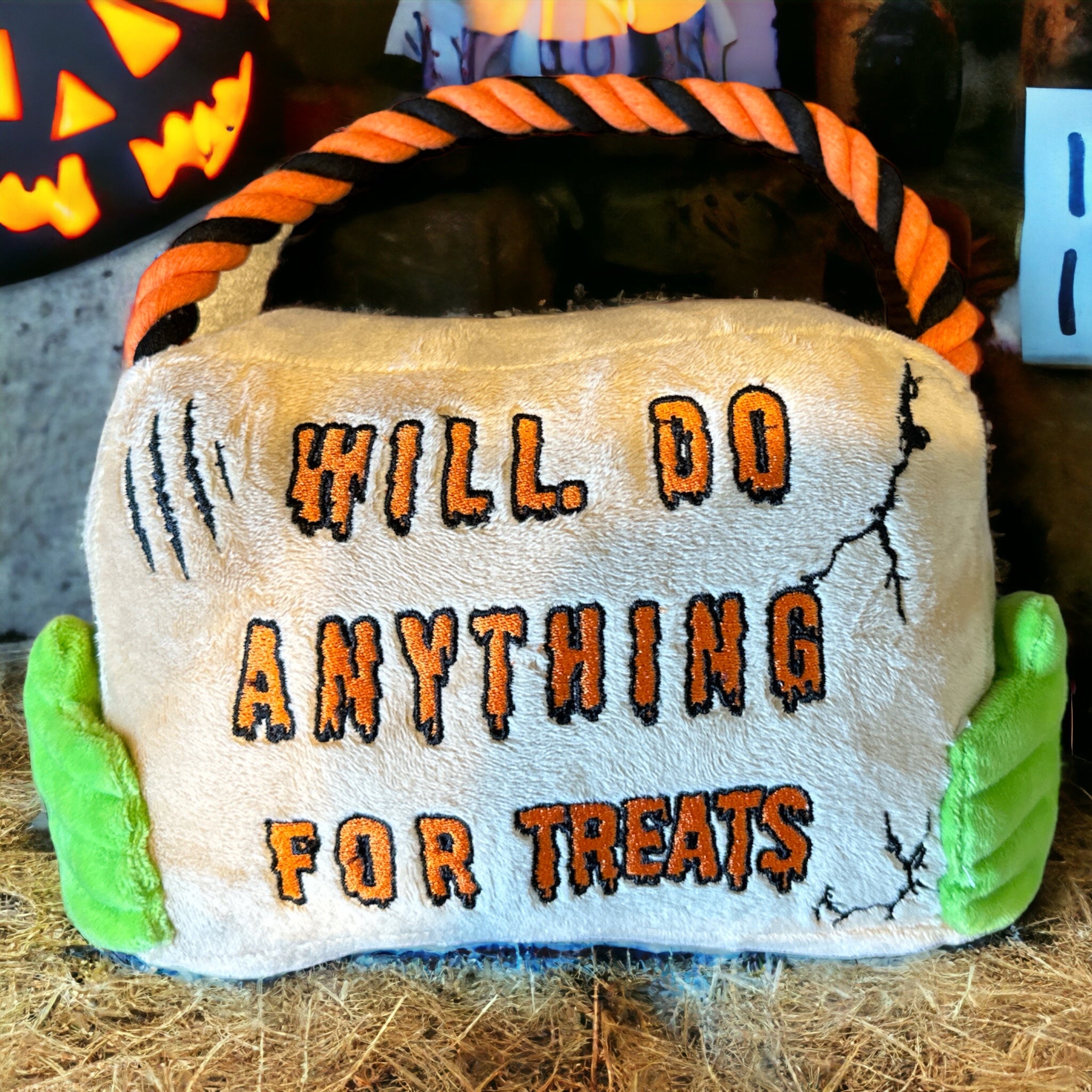 A halloween themed plush dog toy that resembles a sign with a rope. Green hands hold the sign and the sign reads "Will Do Anything For Treats". Set up on a hay bale with pumpkins behind it.