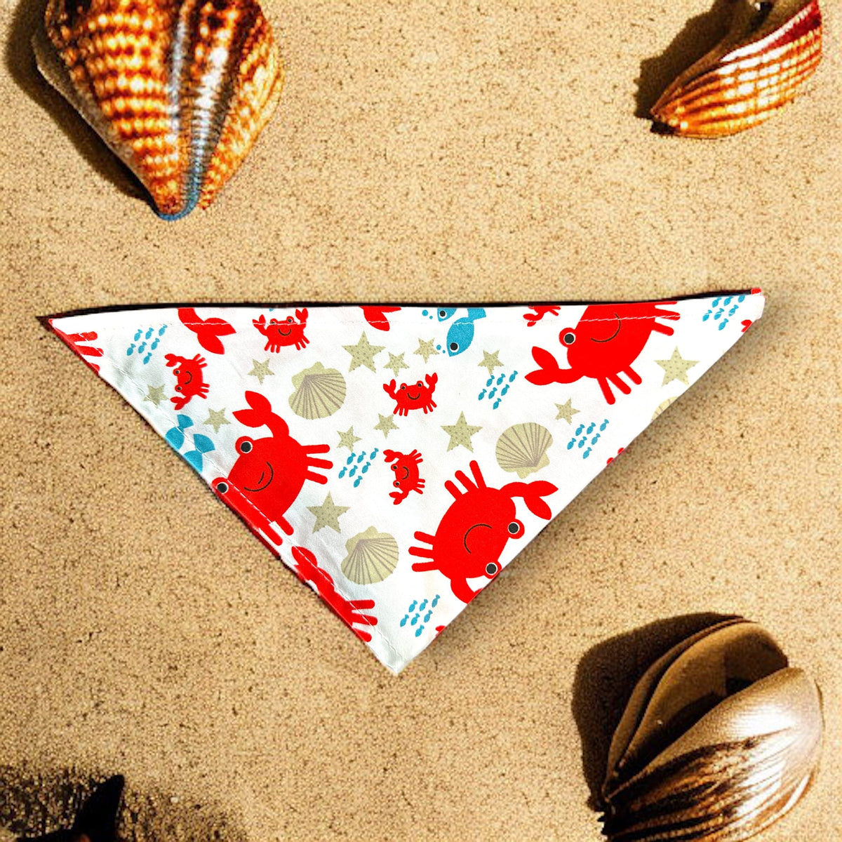 Beach themed dog bandana with images of crabs, shells and fish. The bandana is in the sand with real shells around it. 