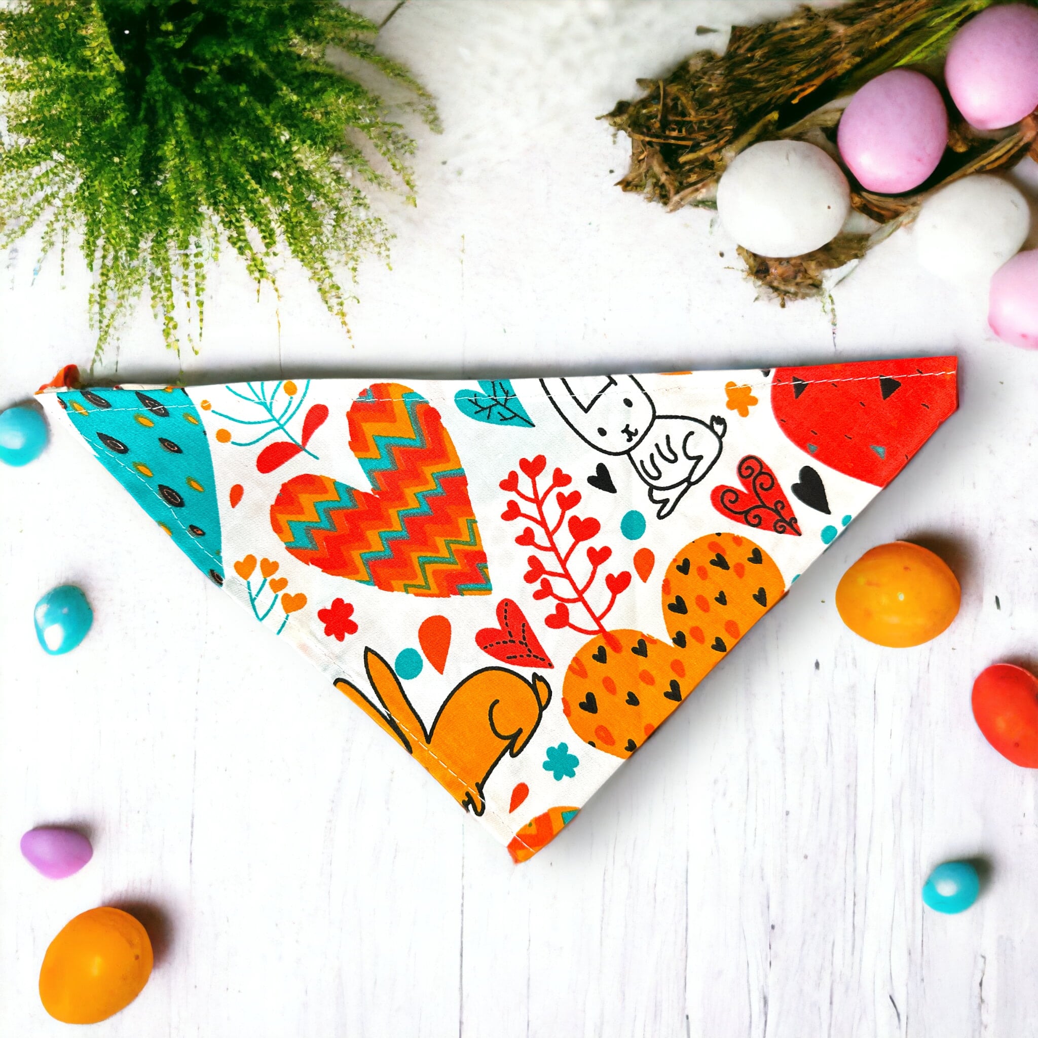 Easter themed dog bandana with colorful easter designs including bunny's, hearts and blue, orange and red colors on a white wooden table with easter eggs and chocolate surrounding.