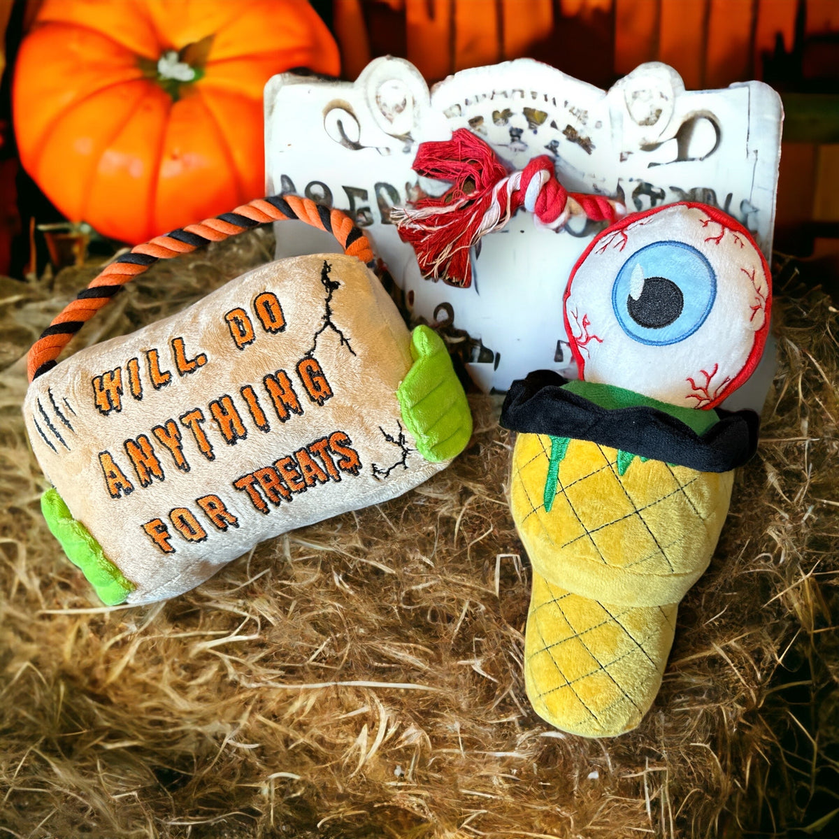 two halloween themed plush dog toys set up on a hay bale with a grave stone and pumpkins in the background. The toy on the left resembles a sign that says &quot;Will Do Anything For Treats&quot; and the toy on the right is an eyeball icecream.